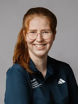 Frau mit roten Haaren, Brille, lächelnd in einem blauen Poloshirt mit einem Adidas-Logo.