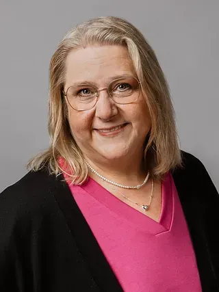Frau mit blonden Haaren, Brille, rosa Hemd und schwarzer Strickjacke lächelt in die Kamera.