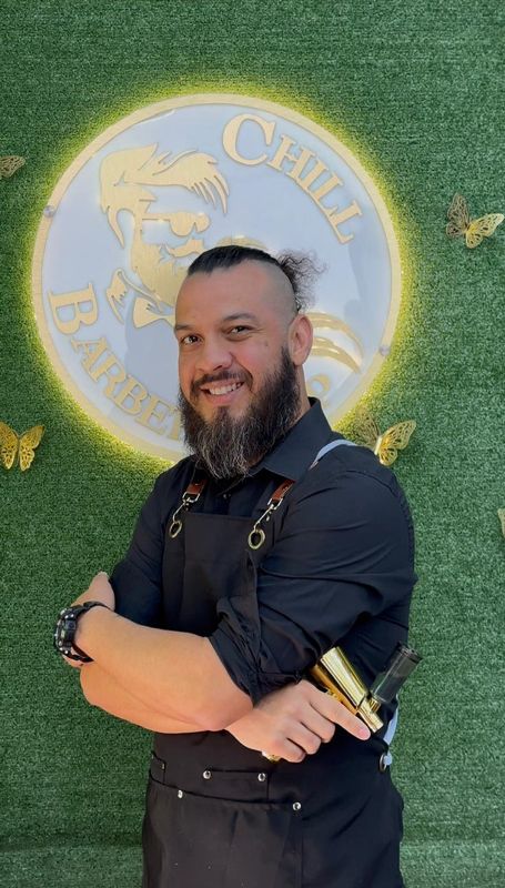 Hombre con barba, brazos cruzados, sostiene una herramienta de oro; logotipo de una barbería en el fondo.