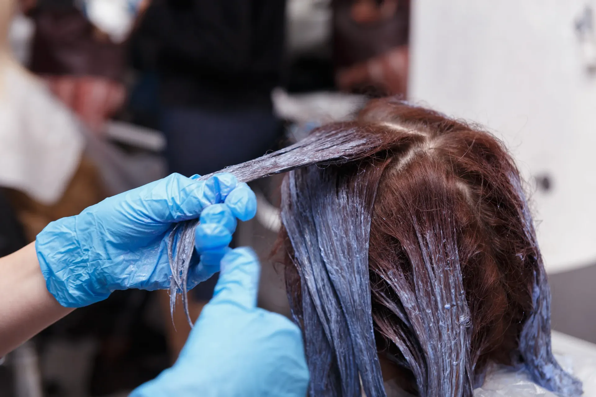 Manos con guantes azules aplicando tinte para el cabello de una persona en un salón.