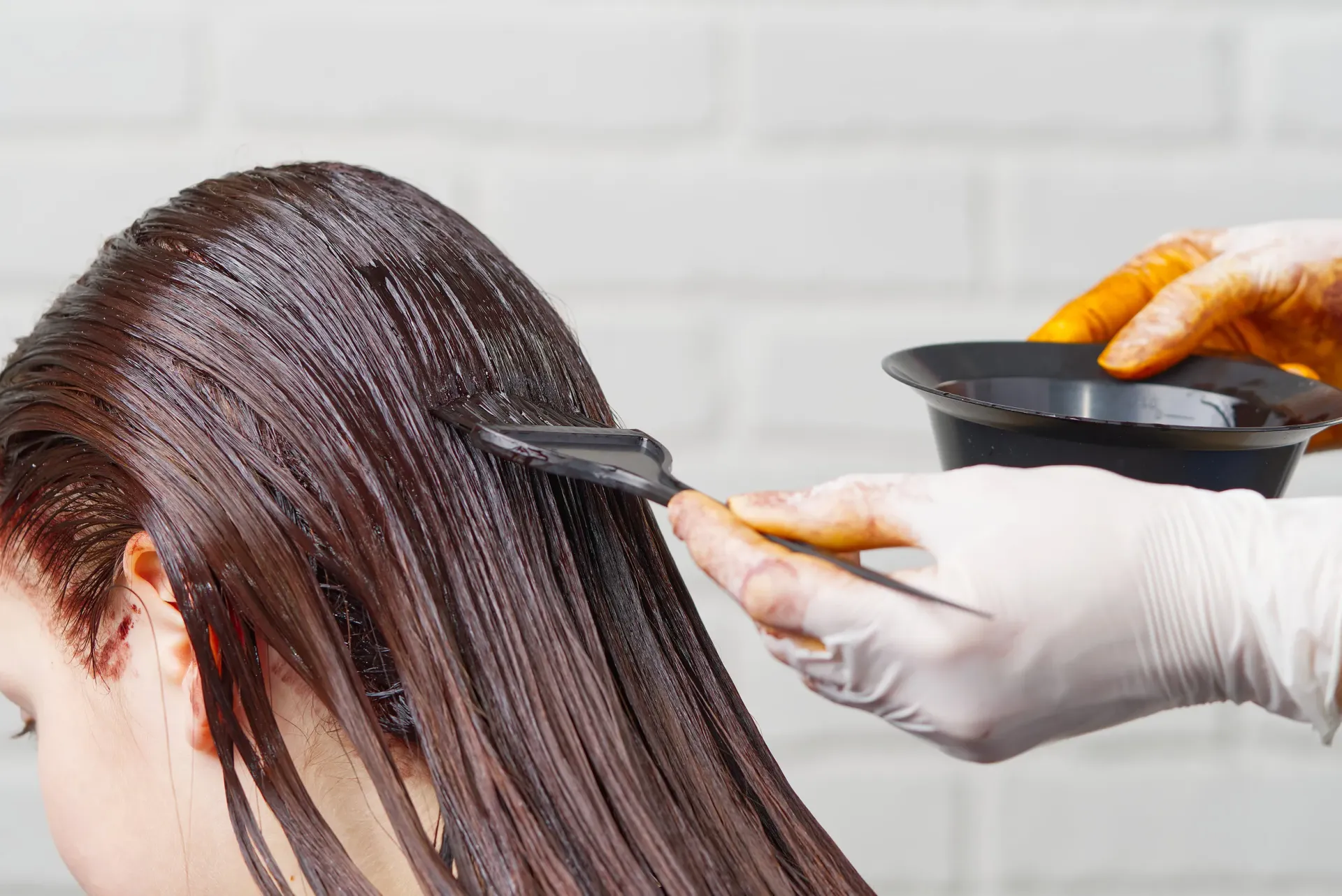 Tinte para el cabello aplicado sobre cabello largo y oscuro con un cepillo, un recipiente y una mano enguantada.