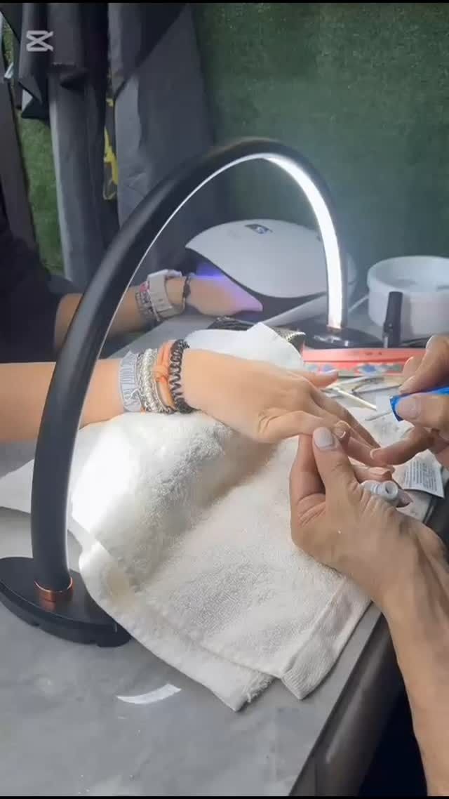 Mano de una persona recibiendo una manicura bajo una lámpara LED curva. Herramientas, toalla y materiales sobre una mesa.