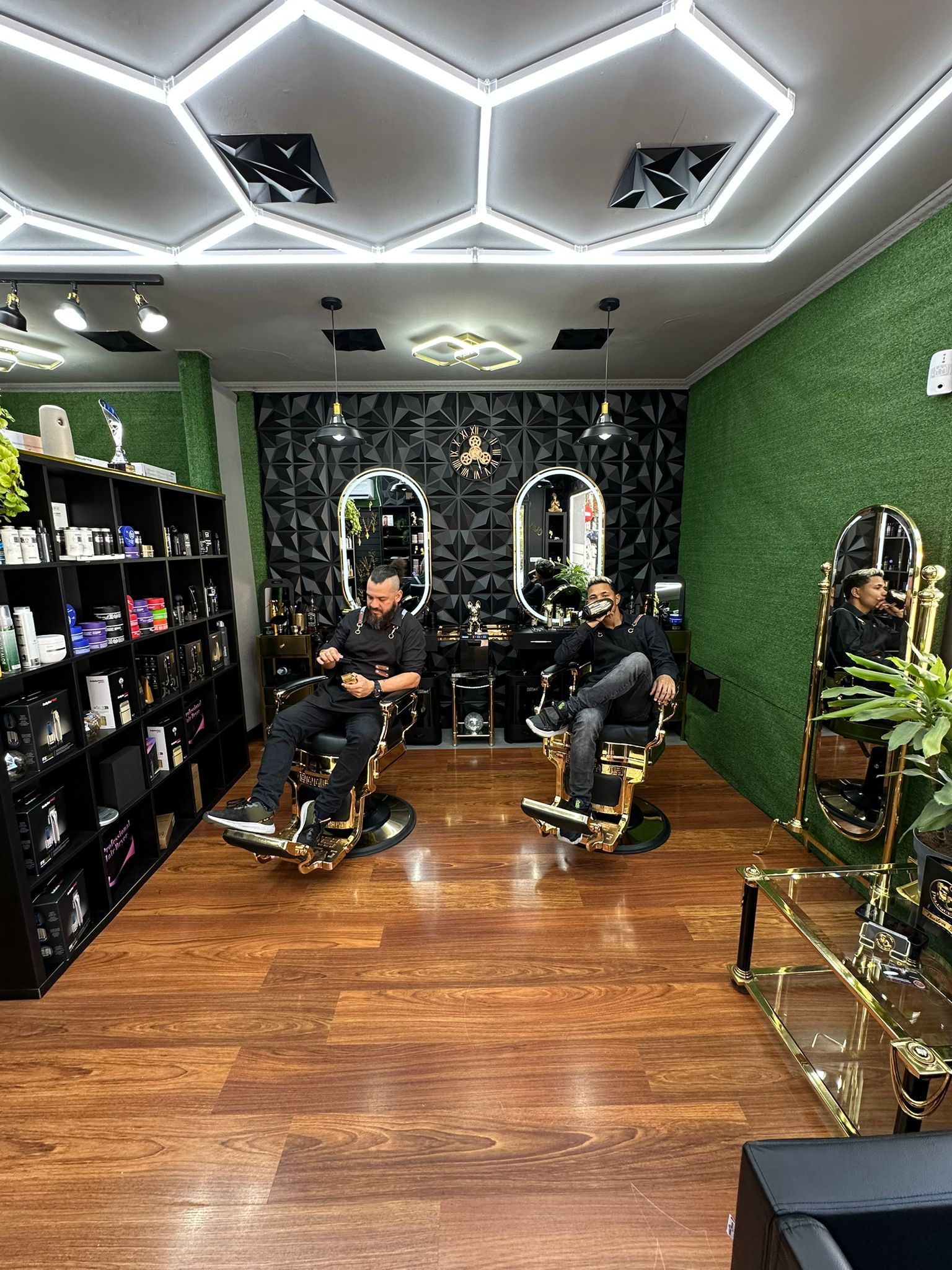Dos hombres sentados en sillones de barbería. El local cuenta con detalles dorados e iluminación decorativa.