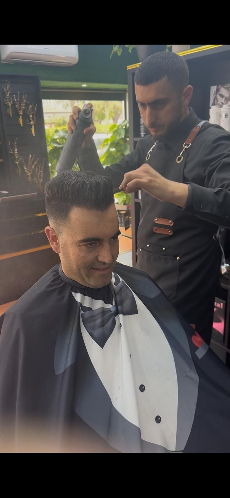 Un barbero corta el pelo a un cliente. El cliente sonríe. El traje del barbero incluye tirantes de cuero y el cliente lleva una capa con diseño de esmoquin.