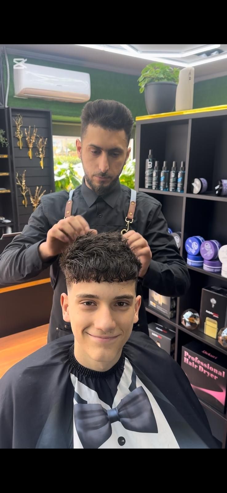 Un barbero le corta el pelo a una clienta en una peluquería. La clienta sonríe. Camisa negra y pajarita.
