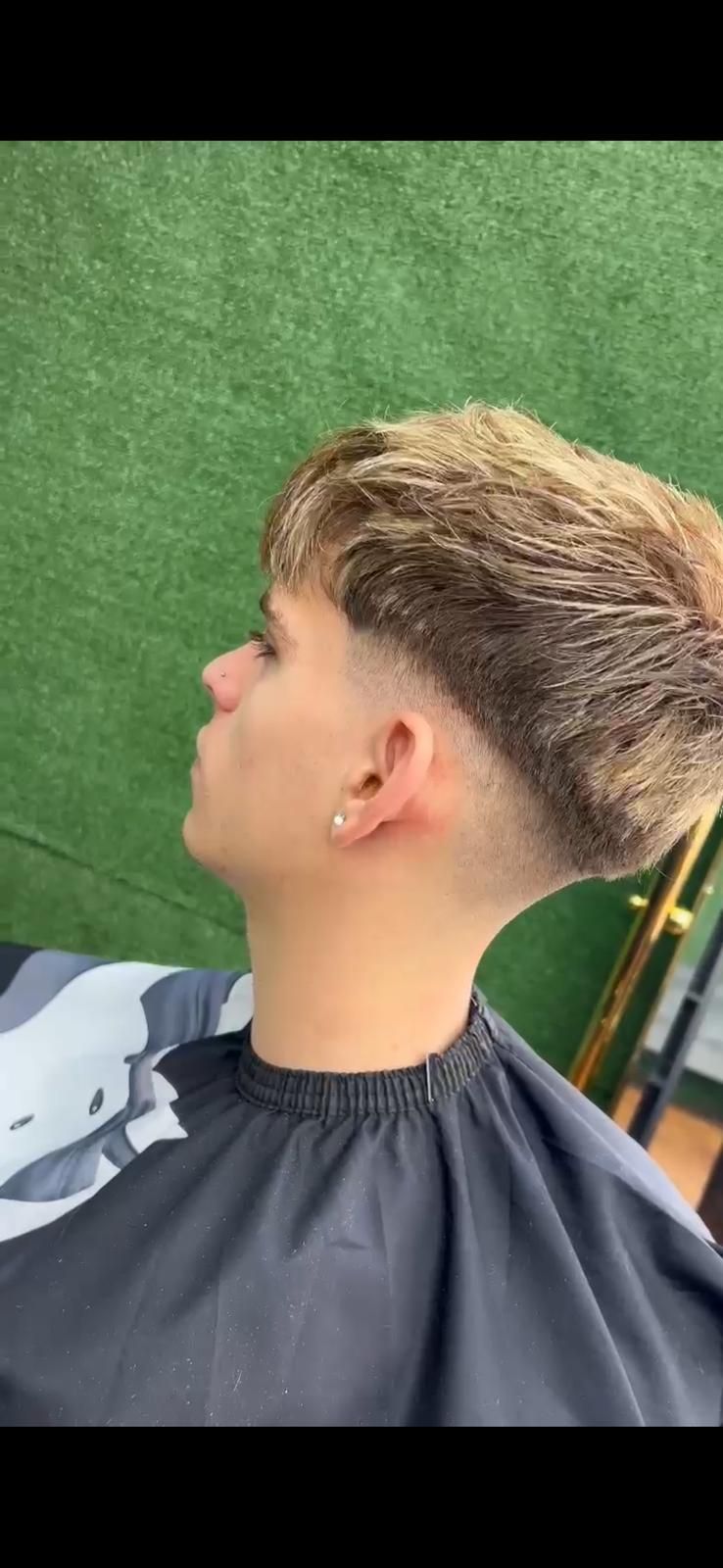 Una persona con un corte de pelo descolorido en una peluquería, con una capa negra. Pared verde al fondo.