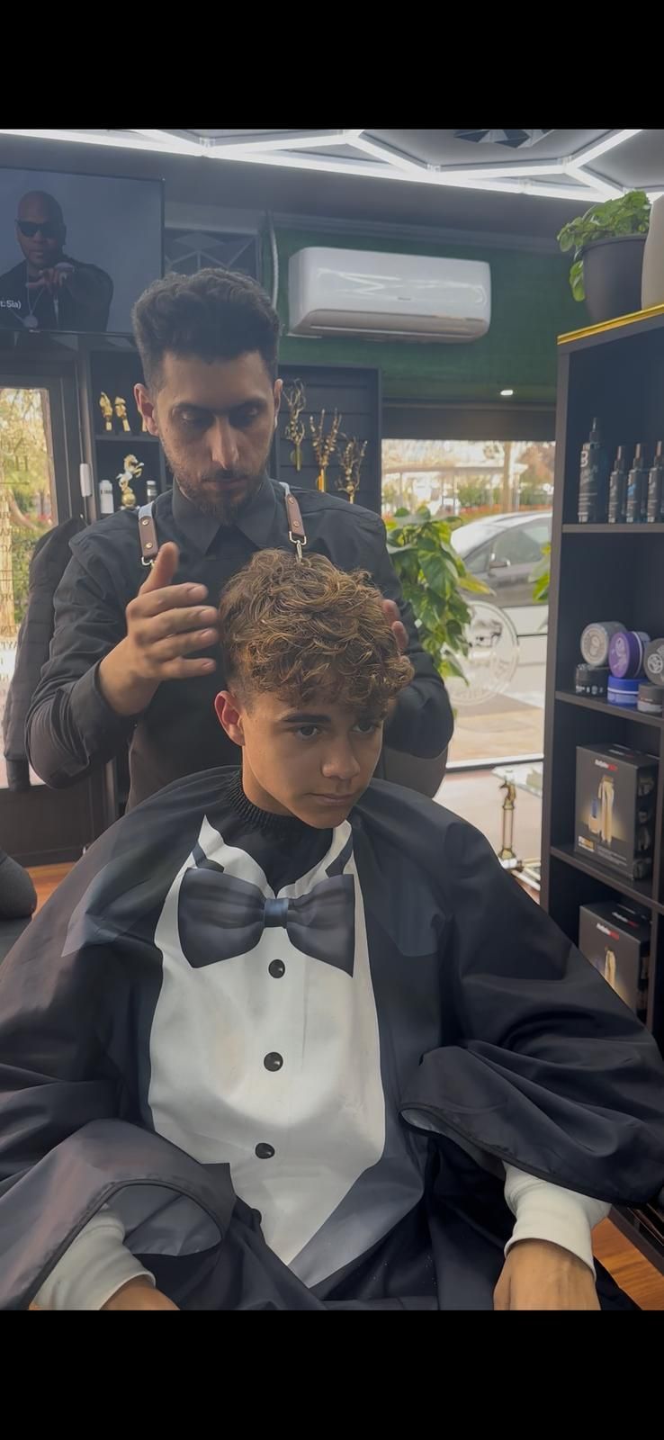 Un barbero corta el pelo rizado de una clienta en un salón. La clienta lleva una capa estilo esmoquin.
