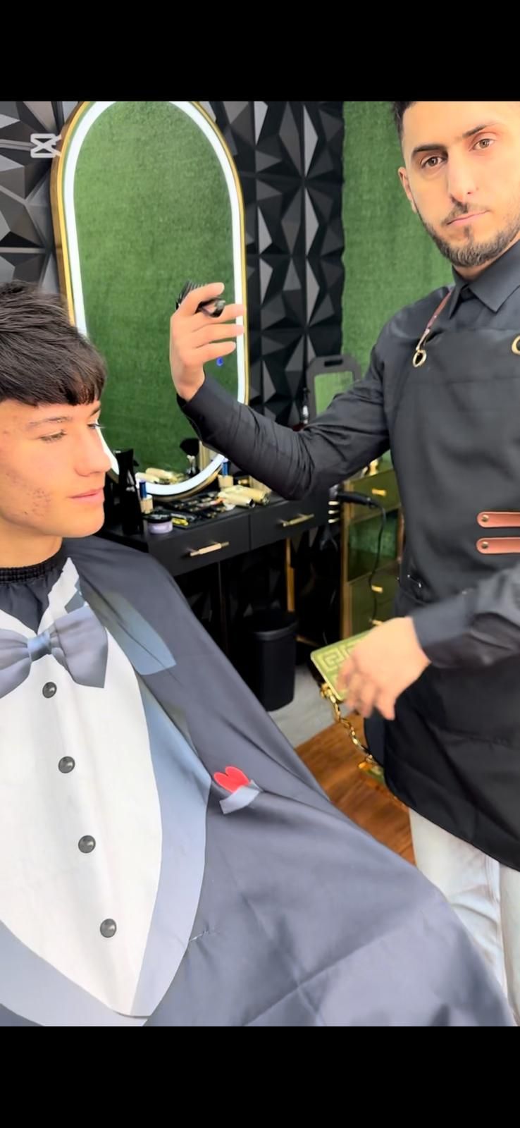 Un barbero corta el pelo a un cliente en un salón. El cliente lleva una capa que parece un esmoquin.