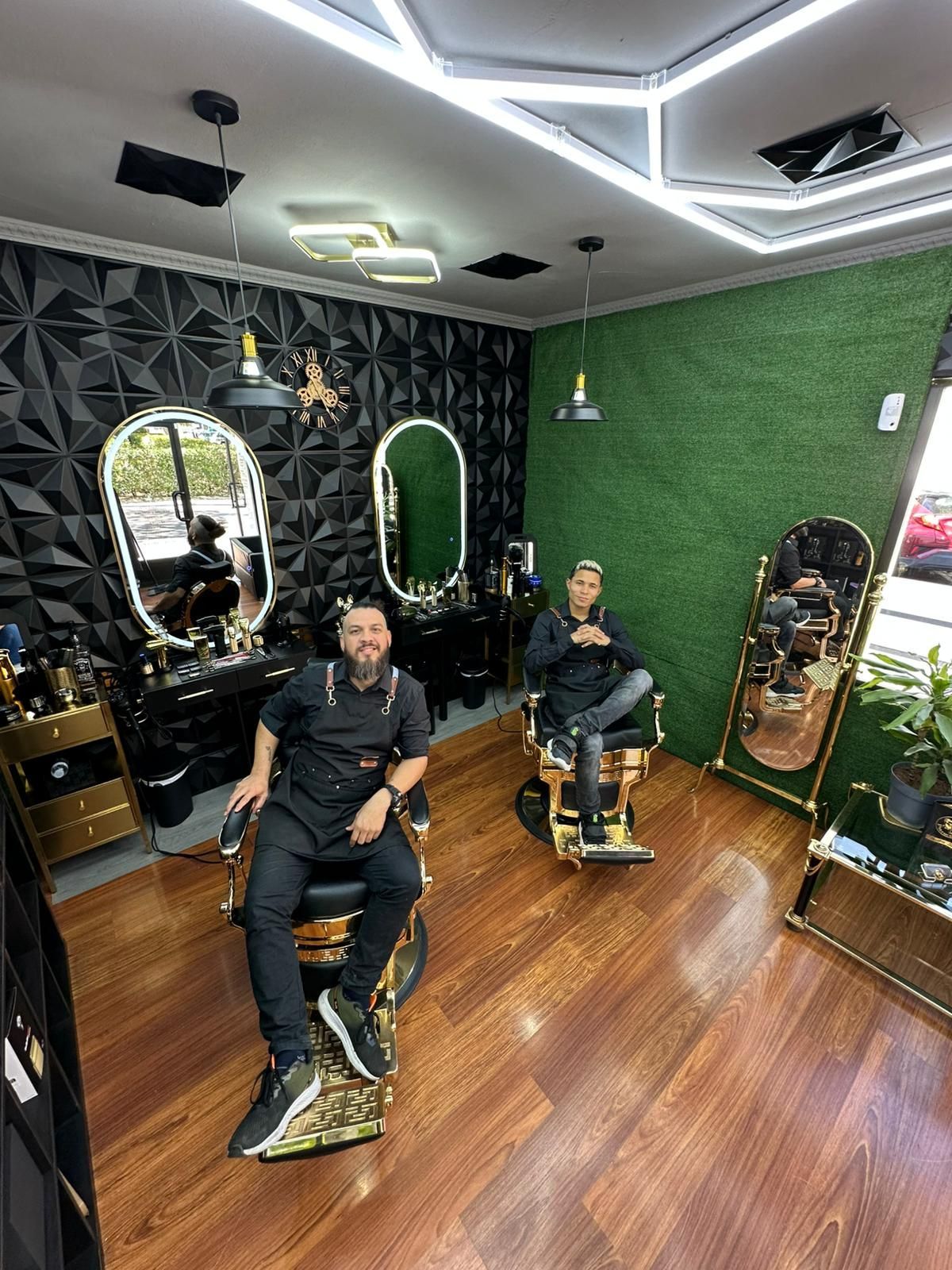 Dos barberos sentados en sillas doradas en una elegante barbería. Uno tiene barba y sonríe.