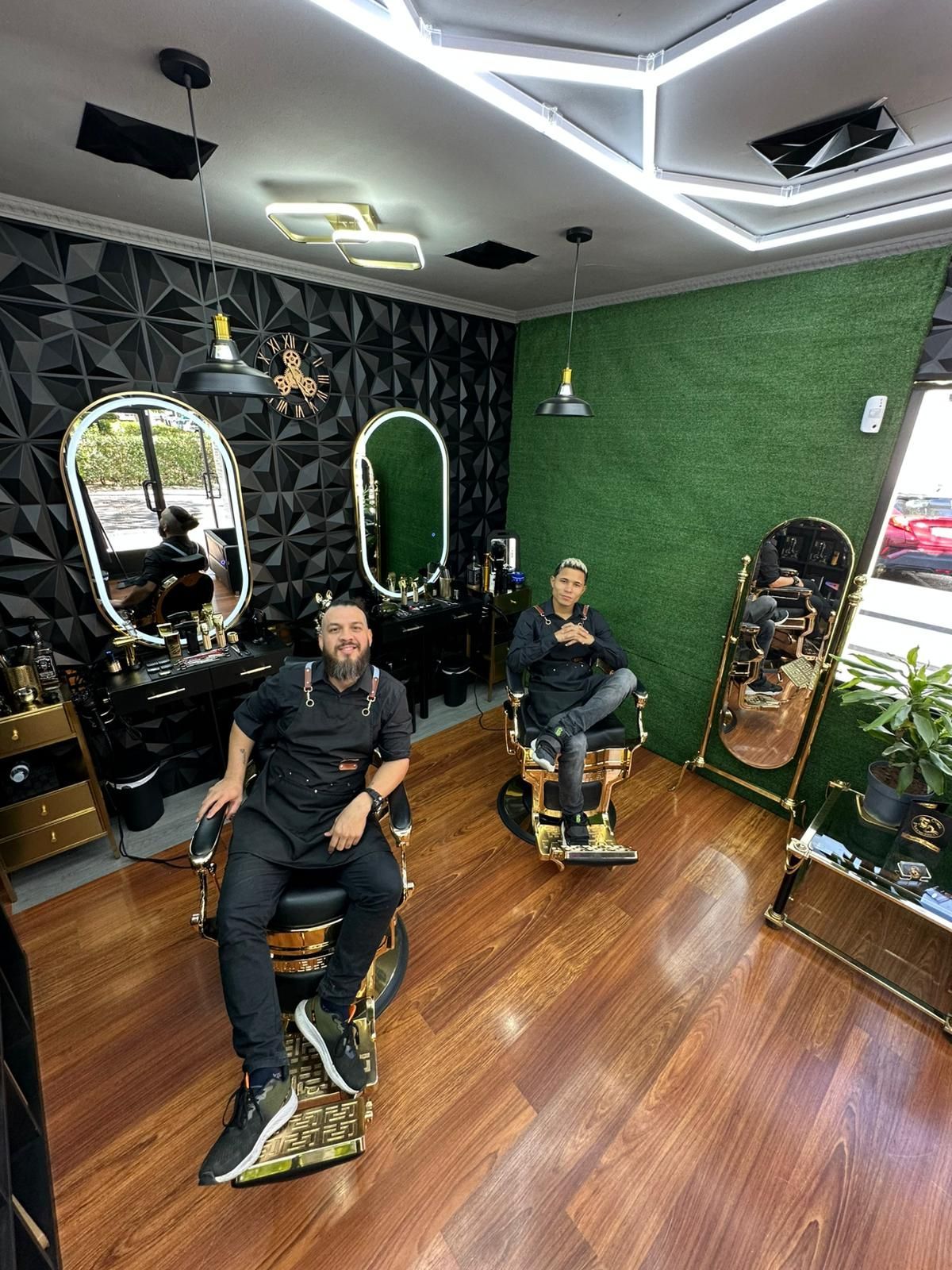 Dos personas sentadas en sillas de barbero. Interior de una barbería con suelos de madera, espejos y paredes negras.