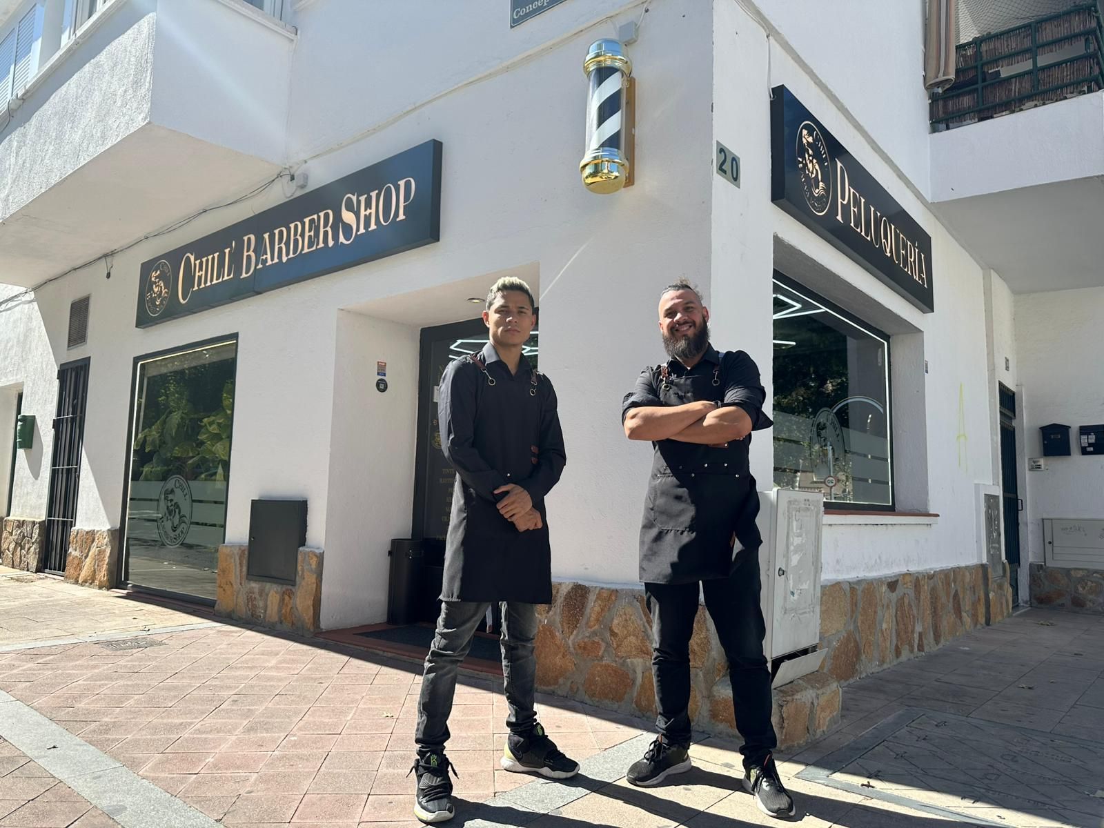 Dos hombres están de pie frente a una barbería con un cartel. Uno tiene los brazos cruzados. El edificio es blanco con detalles en negro.