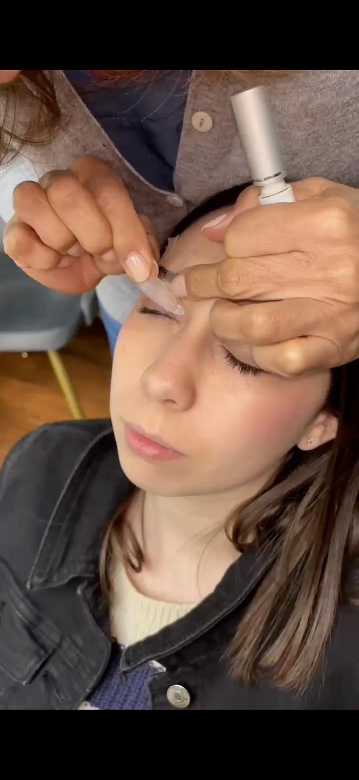 Persona que se está depilando las cejas; sus manos sostienen la tira y el tubo, con la cara hacia arriba.