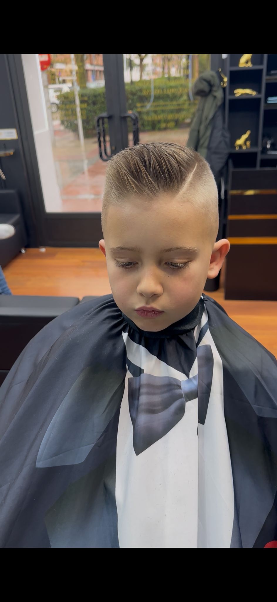 Un joven con un corte de pelo recién cortado sentado en la silla de un barbero, vistiendo una capa con diseño de esmoquin.