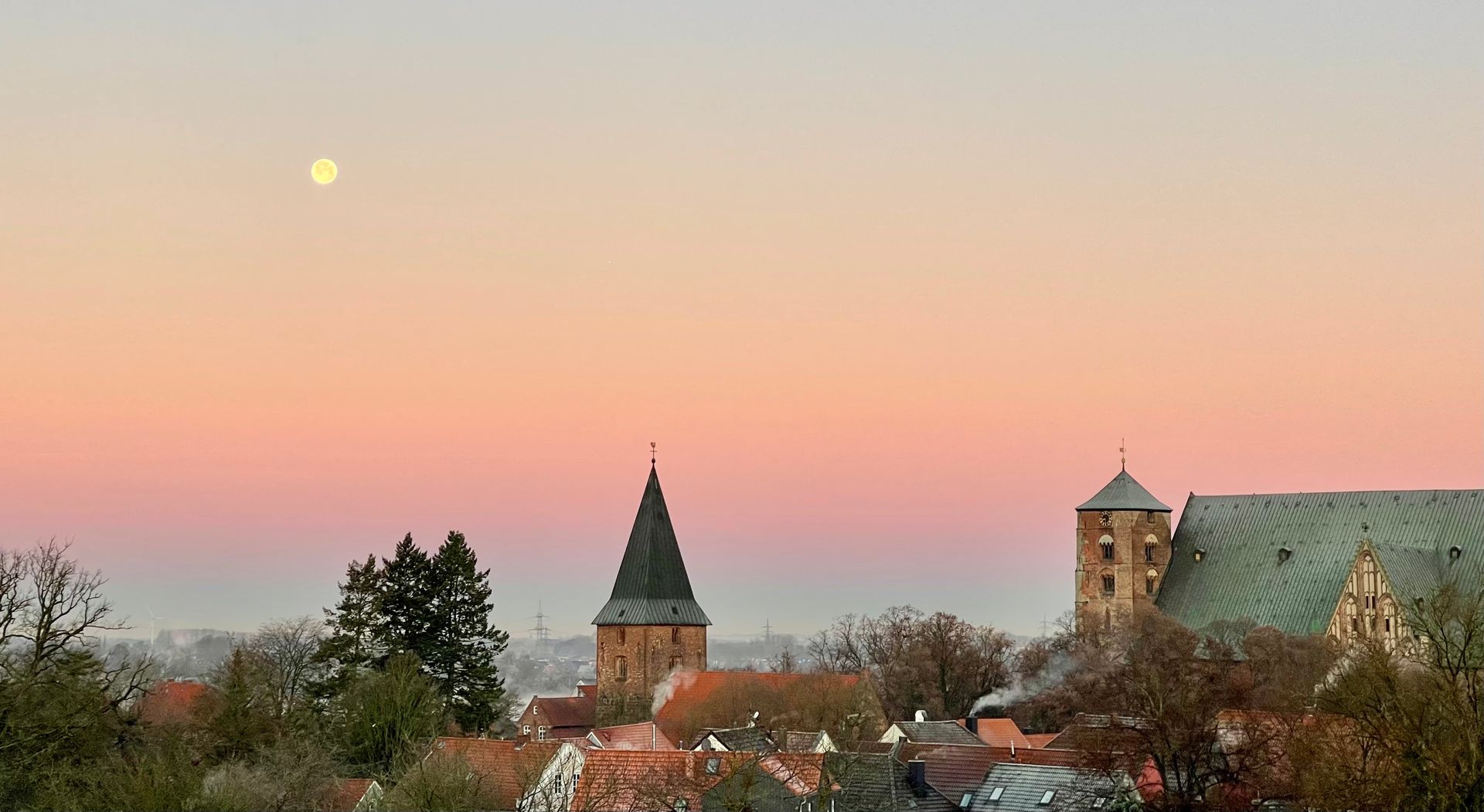 Ein Sonnenuntergang über einer Stadt mit einem Turm im Vordergrund und einem Vollmond im Hintergrund.