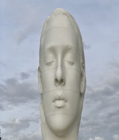 Eine Statue eines Frauengesichts mit geschlossenen Augen vor einem bewölkten Himmel.
