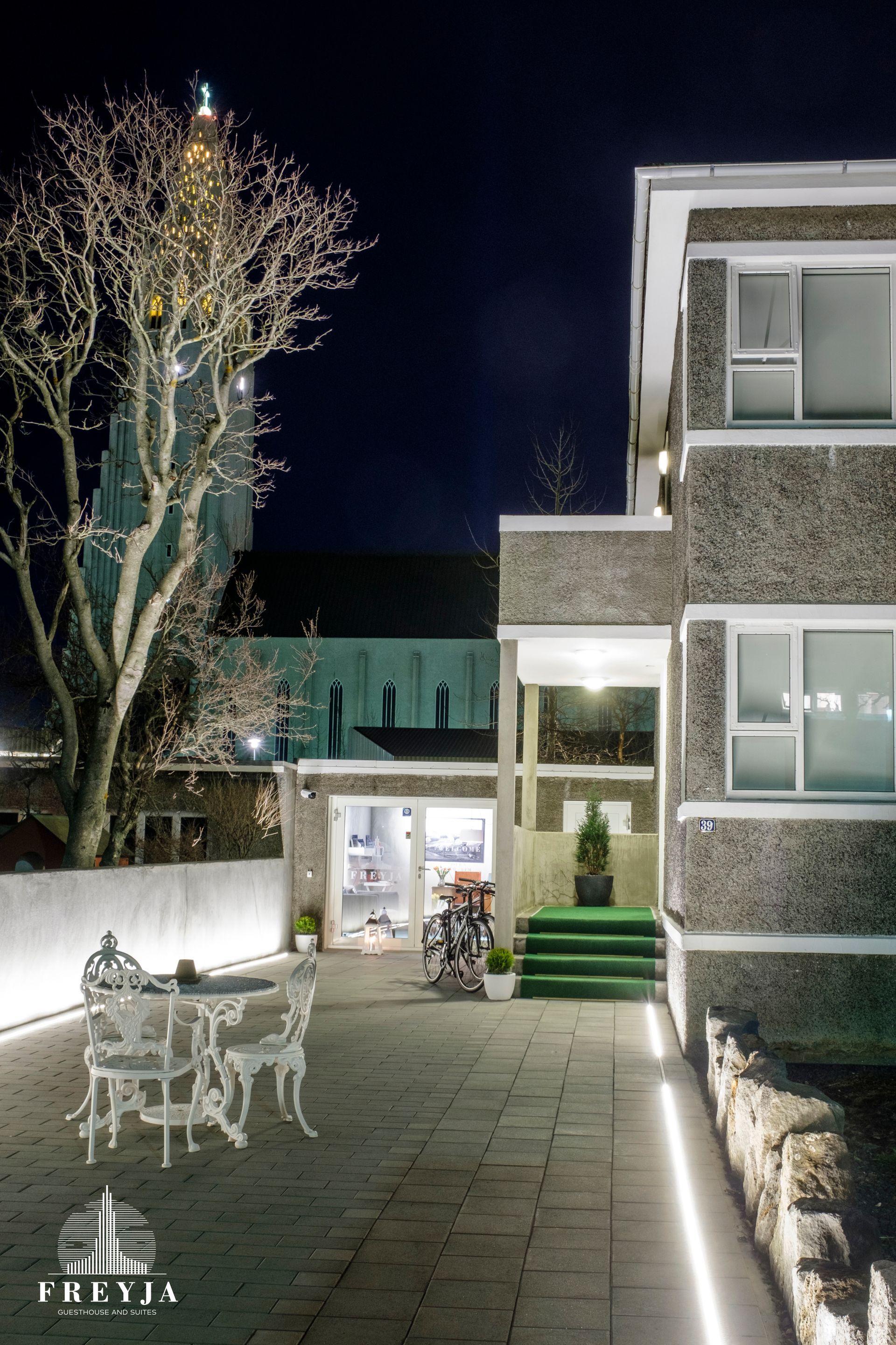 Night shot of a building facade and courtyard with white furniture, with a church in the background.