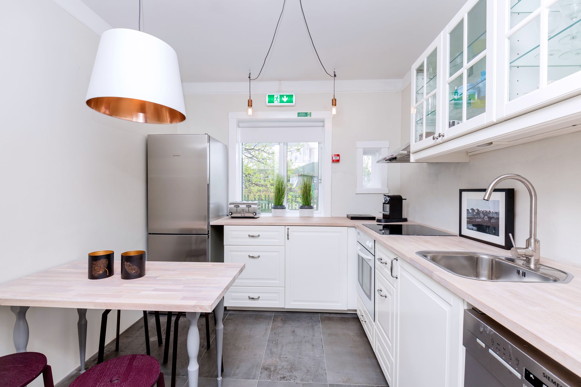 White kitchen with stainless steel fridge, table, and cabinets. Small window, metallic light fixtures.