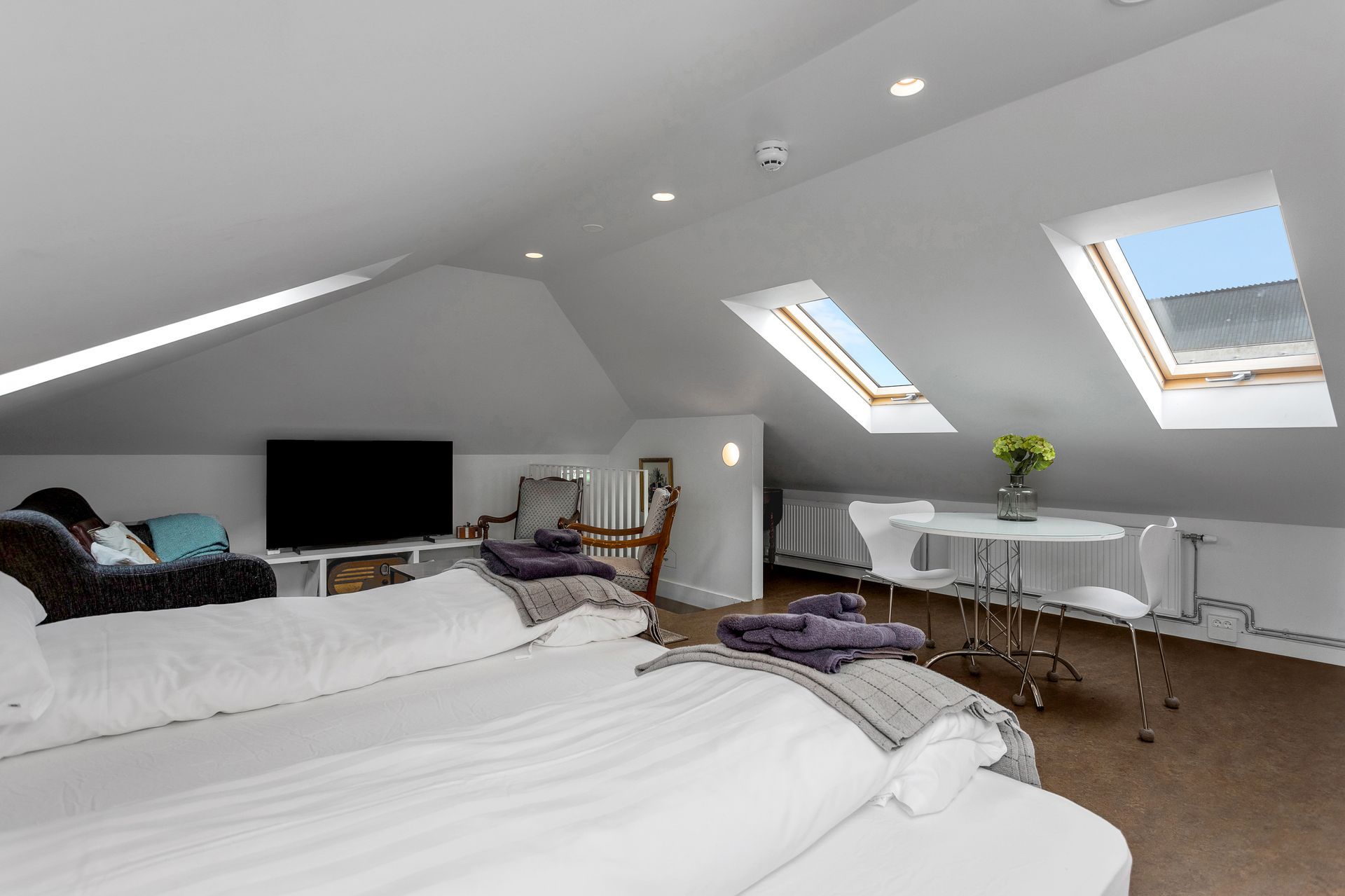 Bedroom with slanted ceiling, skylights, bed, sofa, table, and a large TV. White and light wood interior.