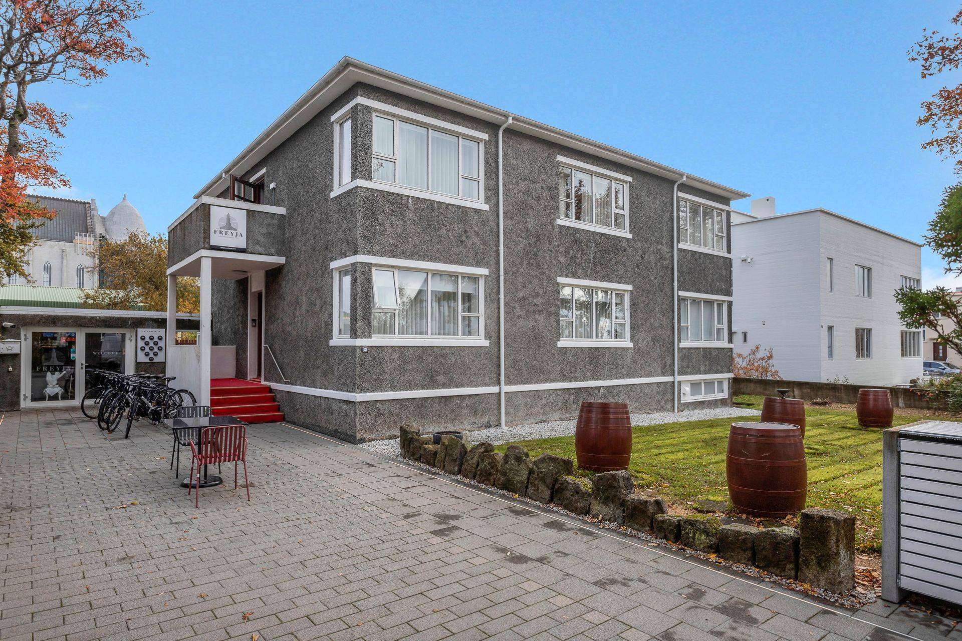Two-story grey building with red steps and white windows. Barrels on the lawn.