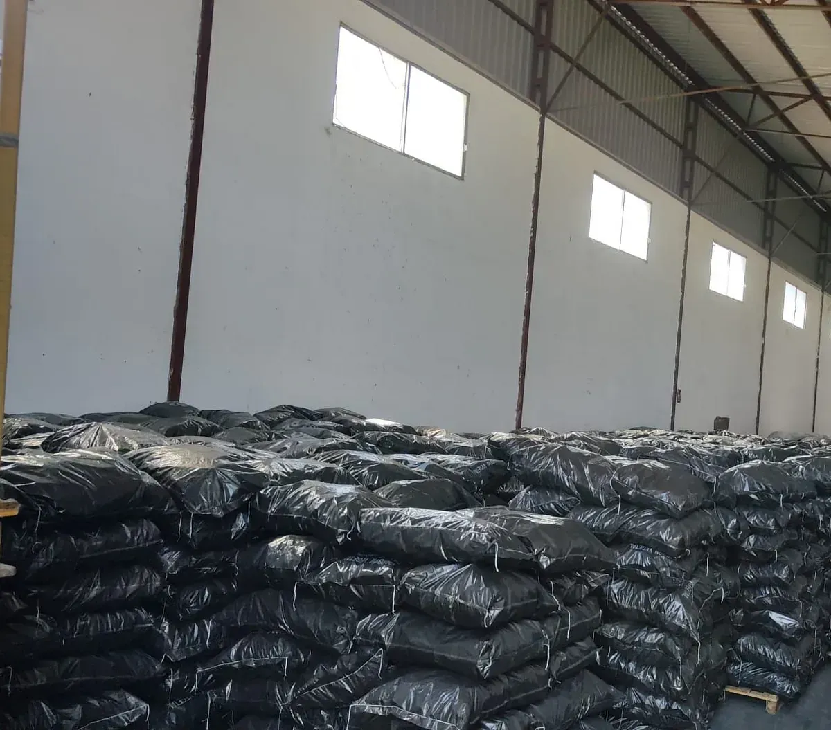 Bolsas de material oscuro apiladas dentro de un almacén con paredes blancas y ventanas altas.