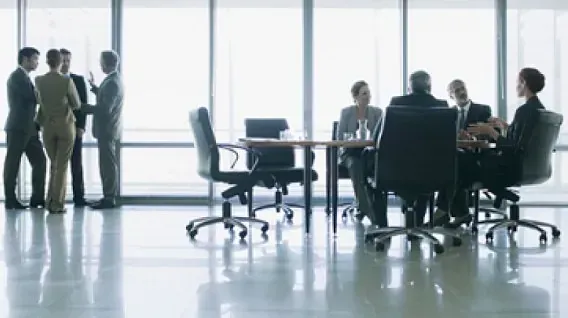 Personas de negocios con traje en una reunión, algunas de pie junto a una ventana, otras sentadas alrededor de una mesa de conferencias.