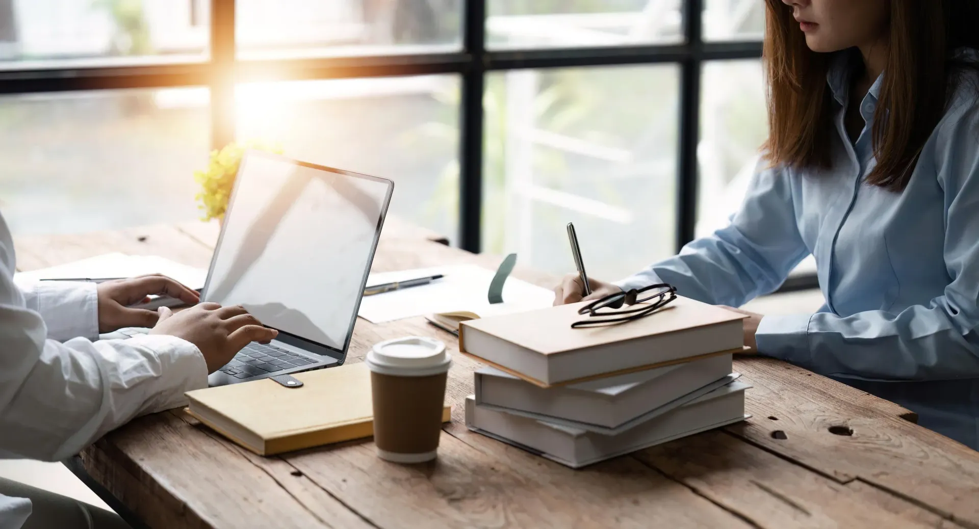 Dos personas trabajan en una mesa de madera: una con una laptop y la otra escribiendo. Hay libros y una taza de café. Luz solar brillante.