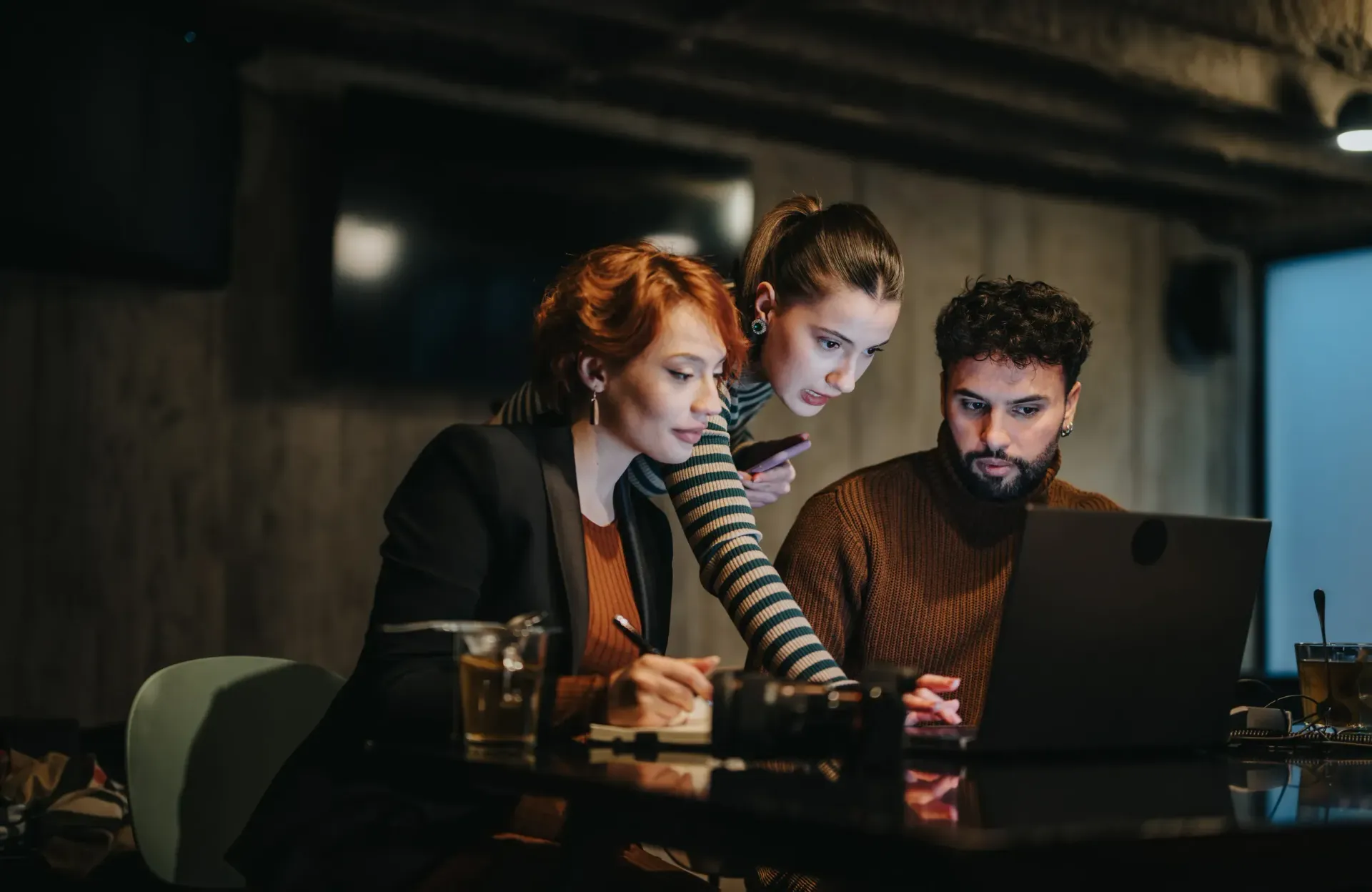 Tres personas mirando una pantalla portátil en un bar poco iluminado, concentrados en un proyecto.