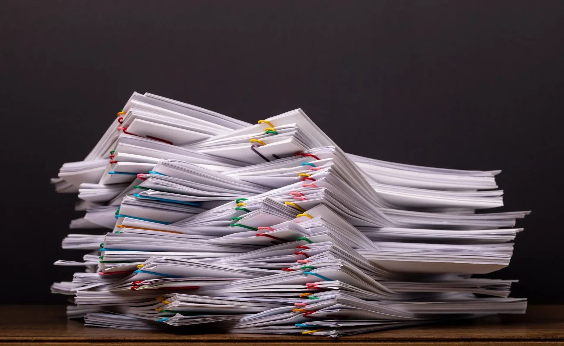 Pila de documentos en papel, algunos unidos con clips, sobre una superficie de madera contra un fondo oscuro.