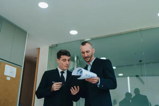 Dos hombres de traje revisando documentos en una oficina, posiblemente discutiendo estrategia.