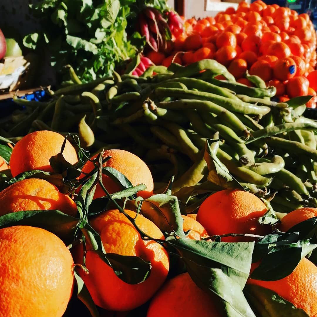 Naranjas, habas y mandarinas apiladas en un puesto de mercado con vegetación y luz solar.
