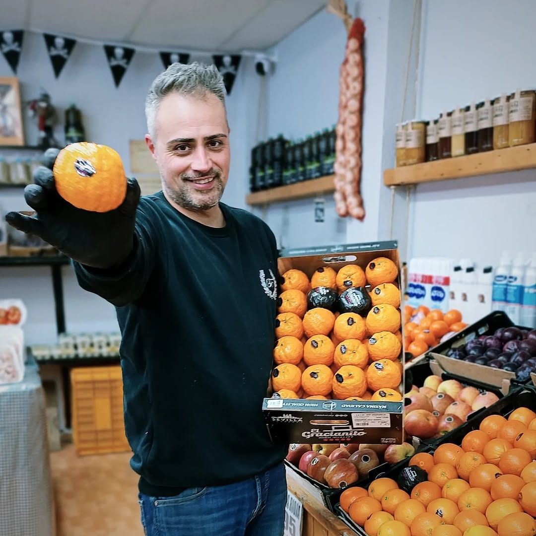 Un hombre en una frutería sostiene una naranja y una caja de naranjas, y sonríe. La tienda tiene frutas y verduras.