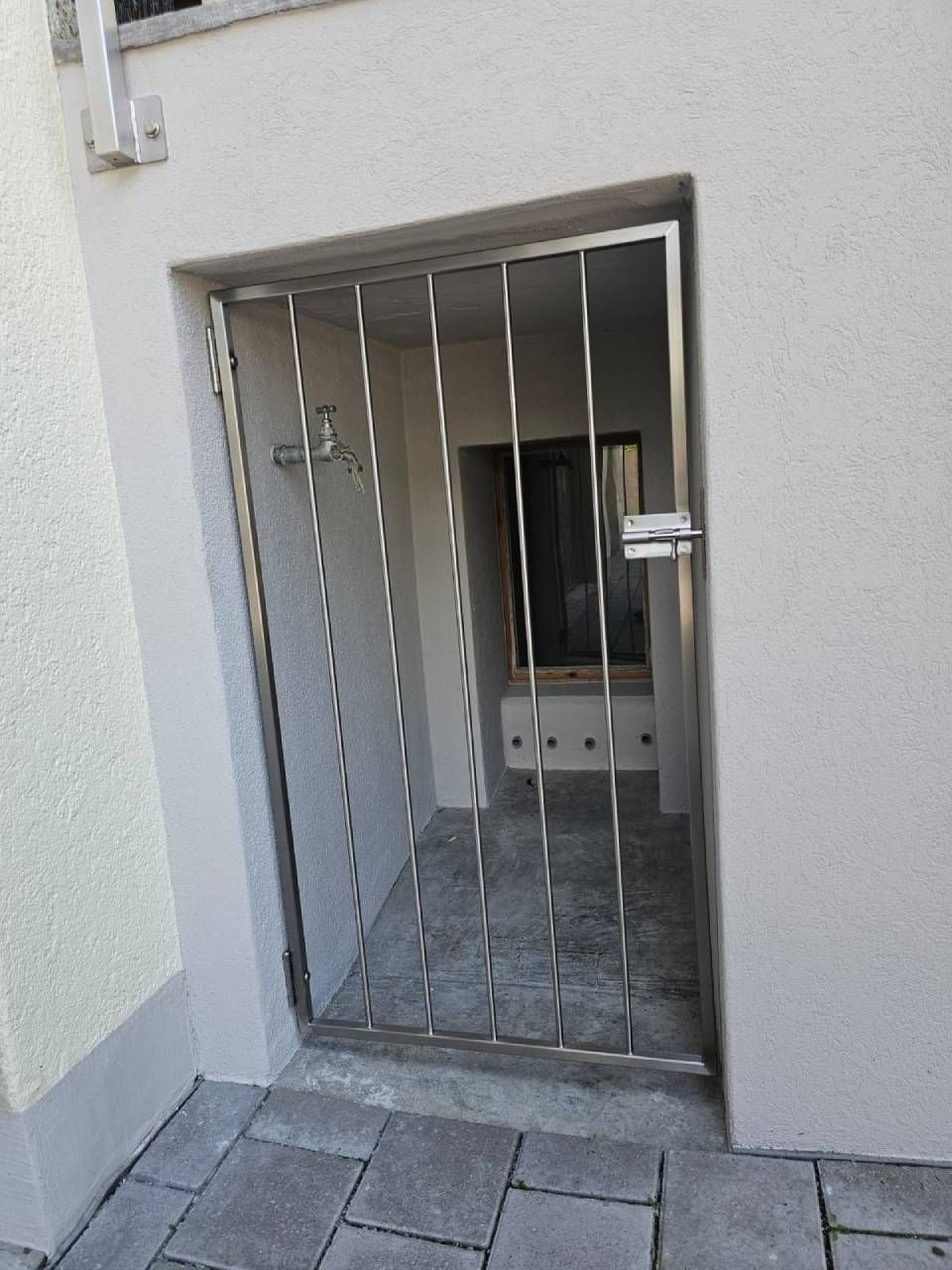In einer Eingangsnische an der hellen Fassade eines Gebäudes befindet sich ein Metalltor mit vertikalen Stäben.