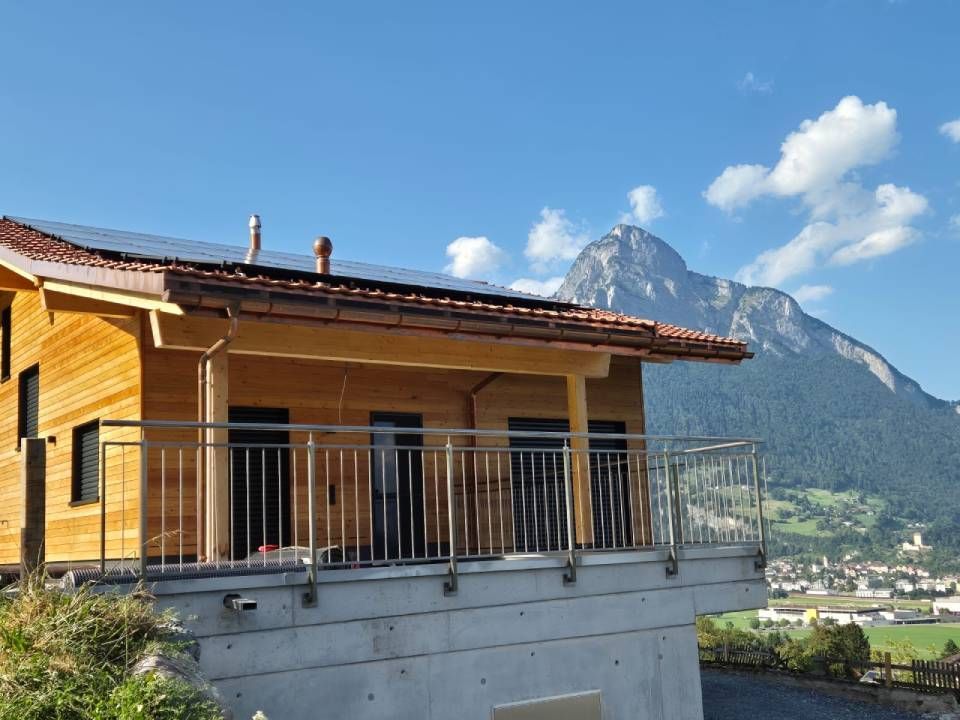Eine moderne Holzhütte mit Metallbalkon steht auf einem Betonsockel vor der Kulisse einer Bergkette und blauem Himmel.