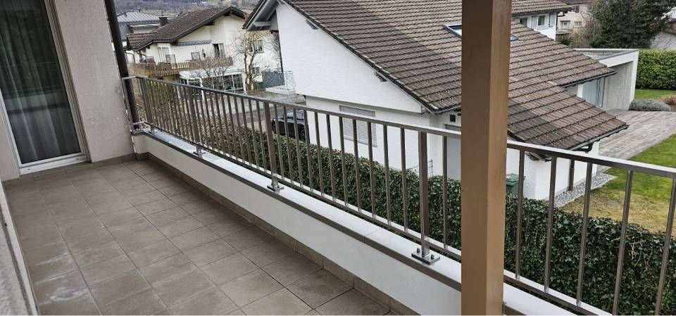 Ein Balkon mit grauem Fliesenboden und Metallgeländer, der auf ein Haus mit braunem Ziegeldach und grüner Hecke blickt.