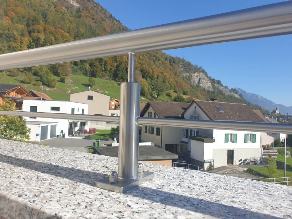 Ein Geländerpfosten aus Edelstahl, montiert an einer Granitwand, mit Blick auf eine Stadt und Bergen im Hintergrund.