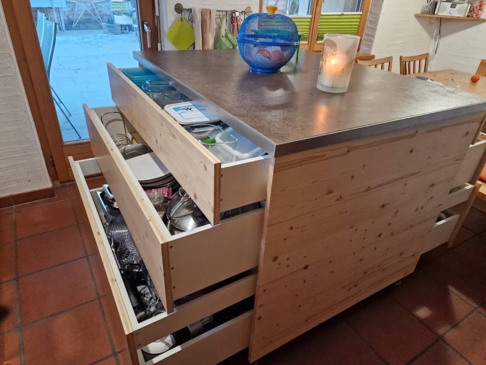 Eine hölzerne Kücheninsel mit drei geöffneten Schubladen, in denen Küchenutensilien aufbewahrt werden, auf einem gefliesten Boden in einem Wohnambiente.