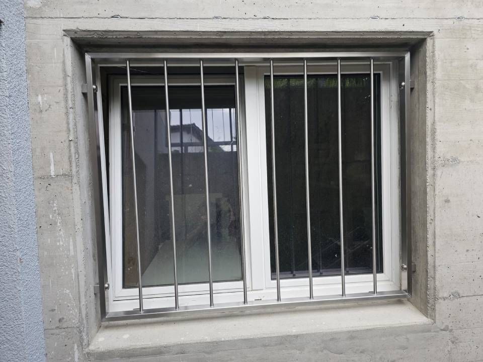 Ein weisser Fensterrahmen in einer Betonwand, abgedeckt von einem vertikalen Sicherheitsgitter aus Metall.