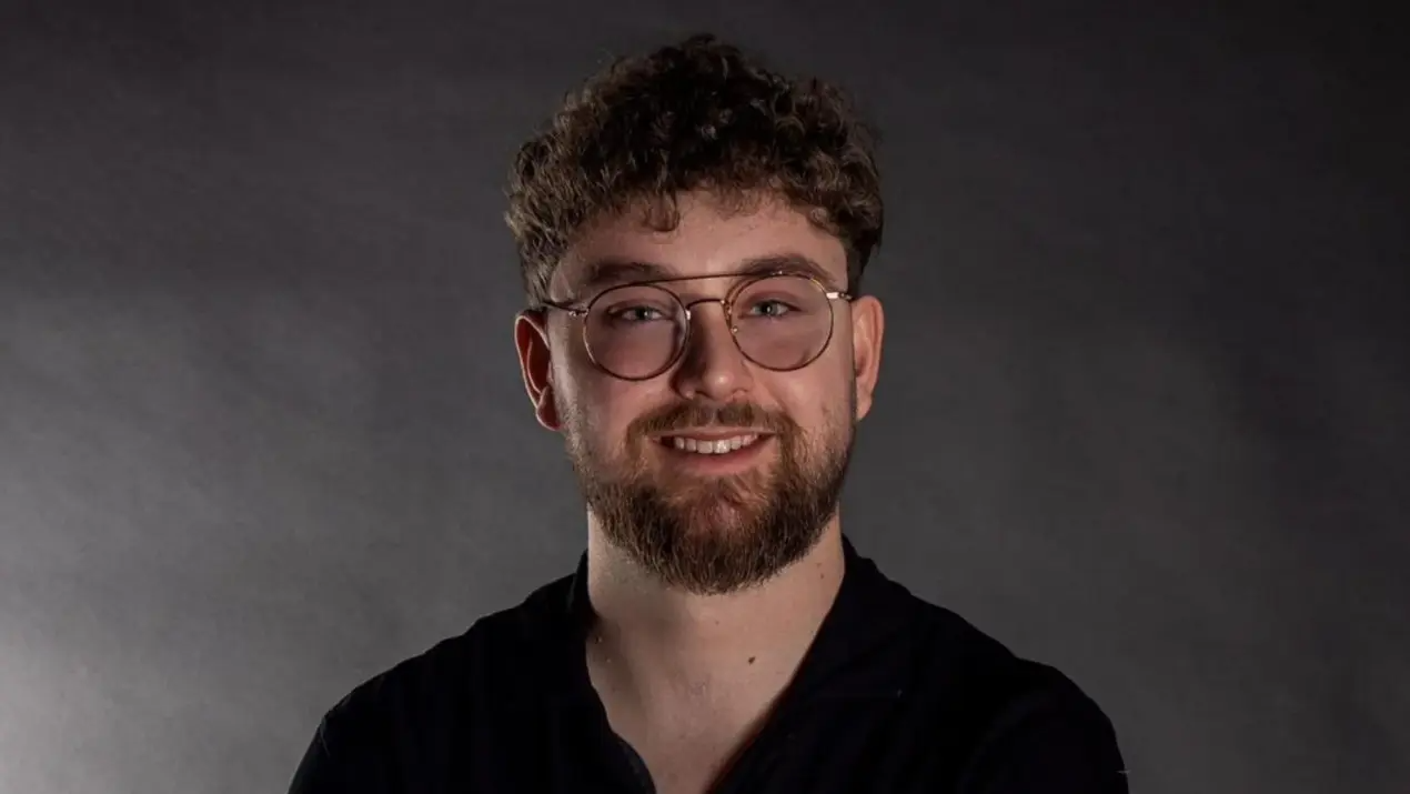 Mann mit Brille und Bart lächelt vor grauem Hintergrund.