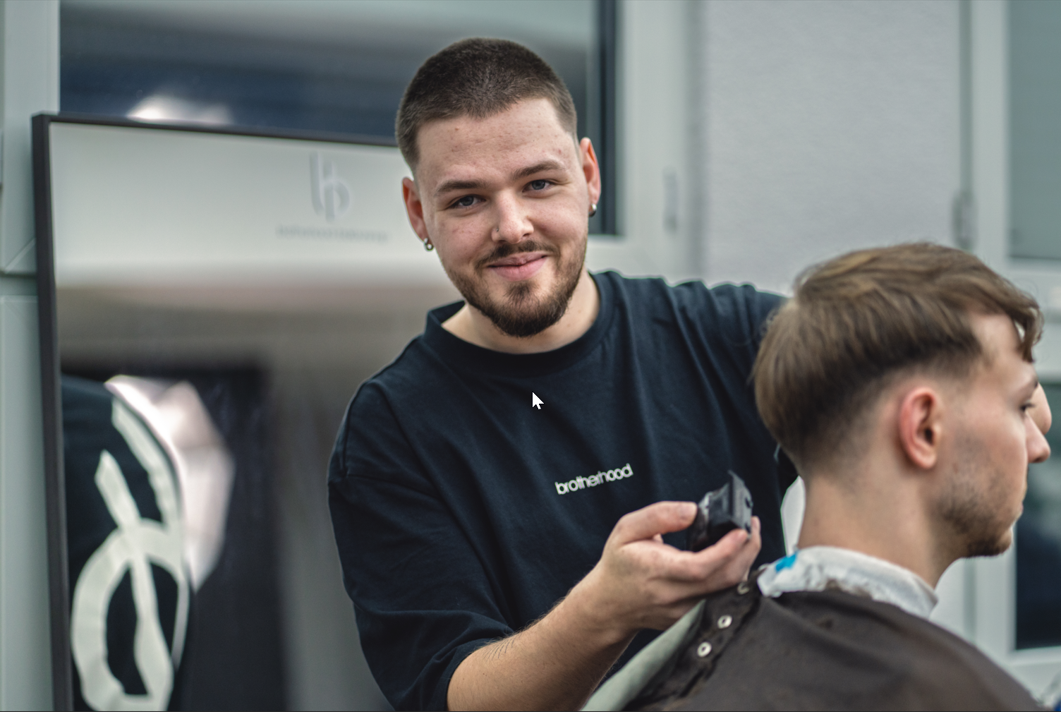 Ein Mann im grünen Pullover blickt in die Kamera. Er steht in einem Flur; im Hintergrund sind zwei gerahmte Kunstwerke und eine Lampe zu sehen.