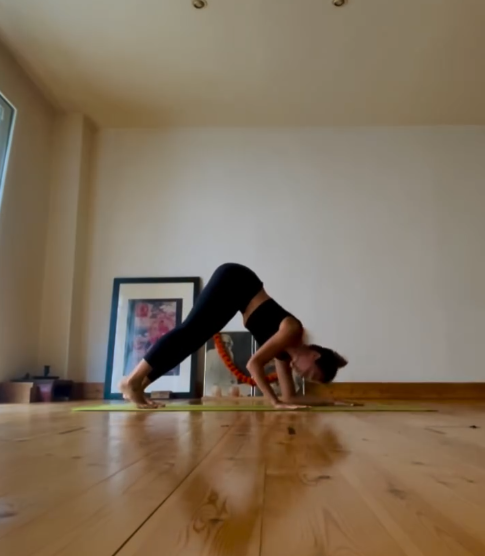 Una mujer vestida con ropa deportiva negra realiza una postura de yoga con la cabeza cerca del suelo, sobre una esterilla.
