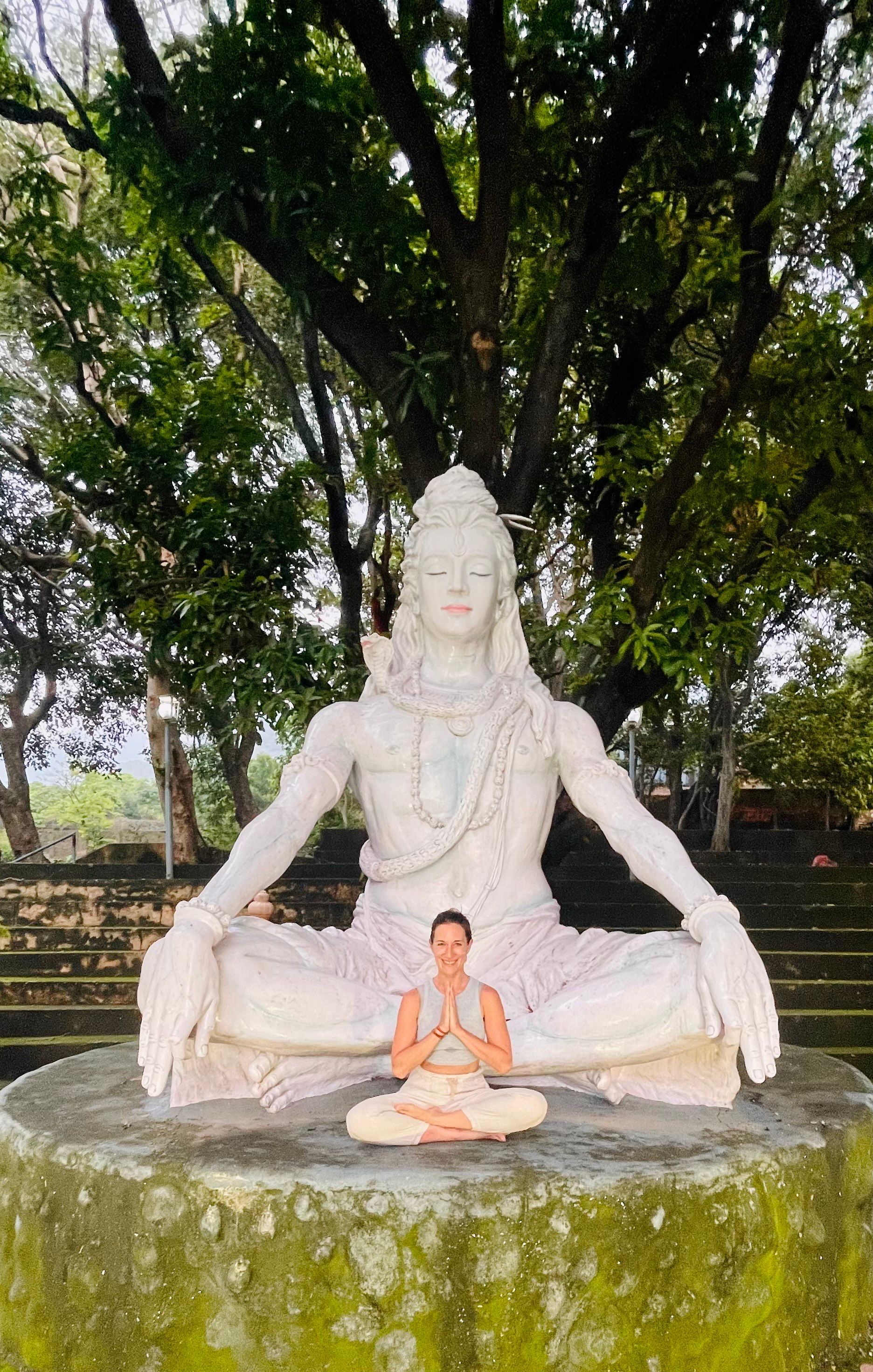 Persona meditando frente a una estatua blanca de Shiva al aire libre, sentada sobre una plataforma de piedra.