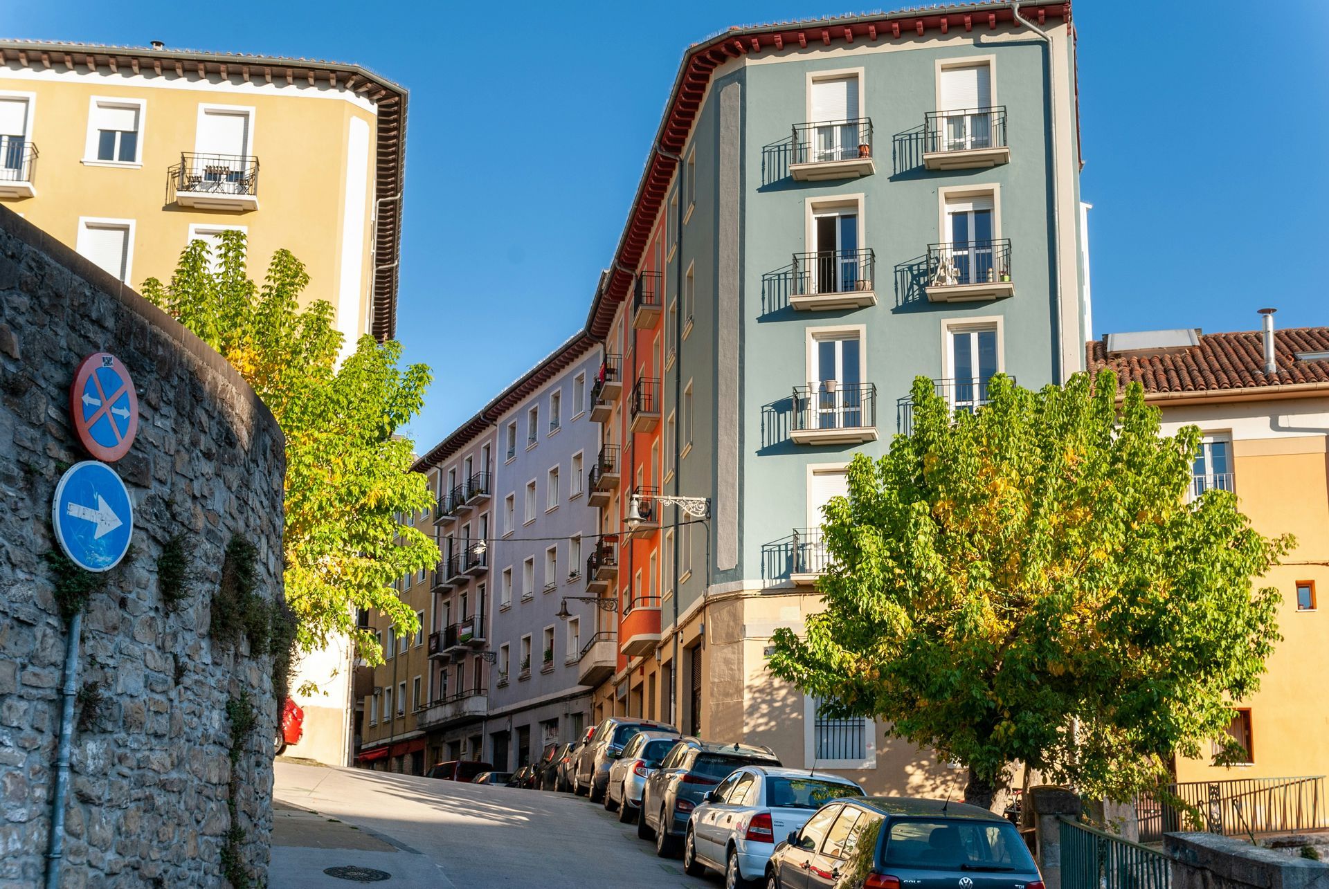 Una estrecha calle europea con edificios coloridos, coches aparcados y un árbol, bajo un cielo azul claro.