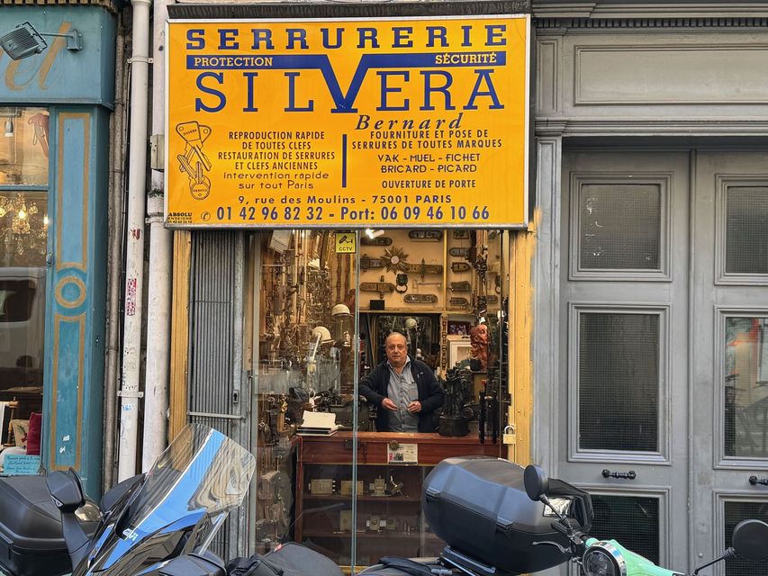 La façade d'un magsin de serrurier avec une enseigne jaune indiquant « Serrurerie Silvera » au-dessus d'un homme debout derrière un comptoir vitré.