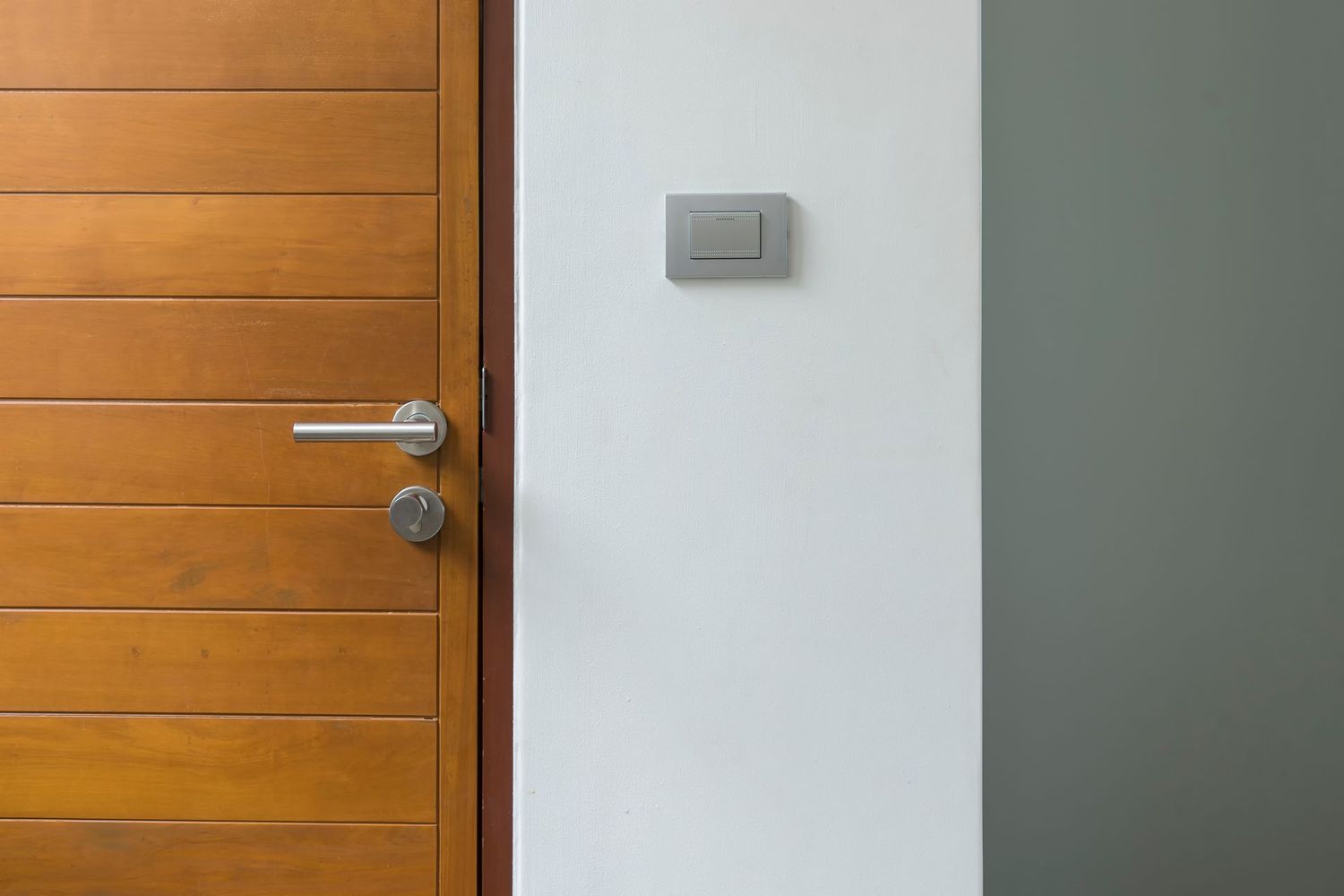 Une porte en bois avec une poignée et une serrure argentées modernes, adossée à un mur blanc où se trouve un petit interrupteur carré.
