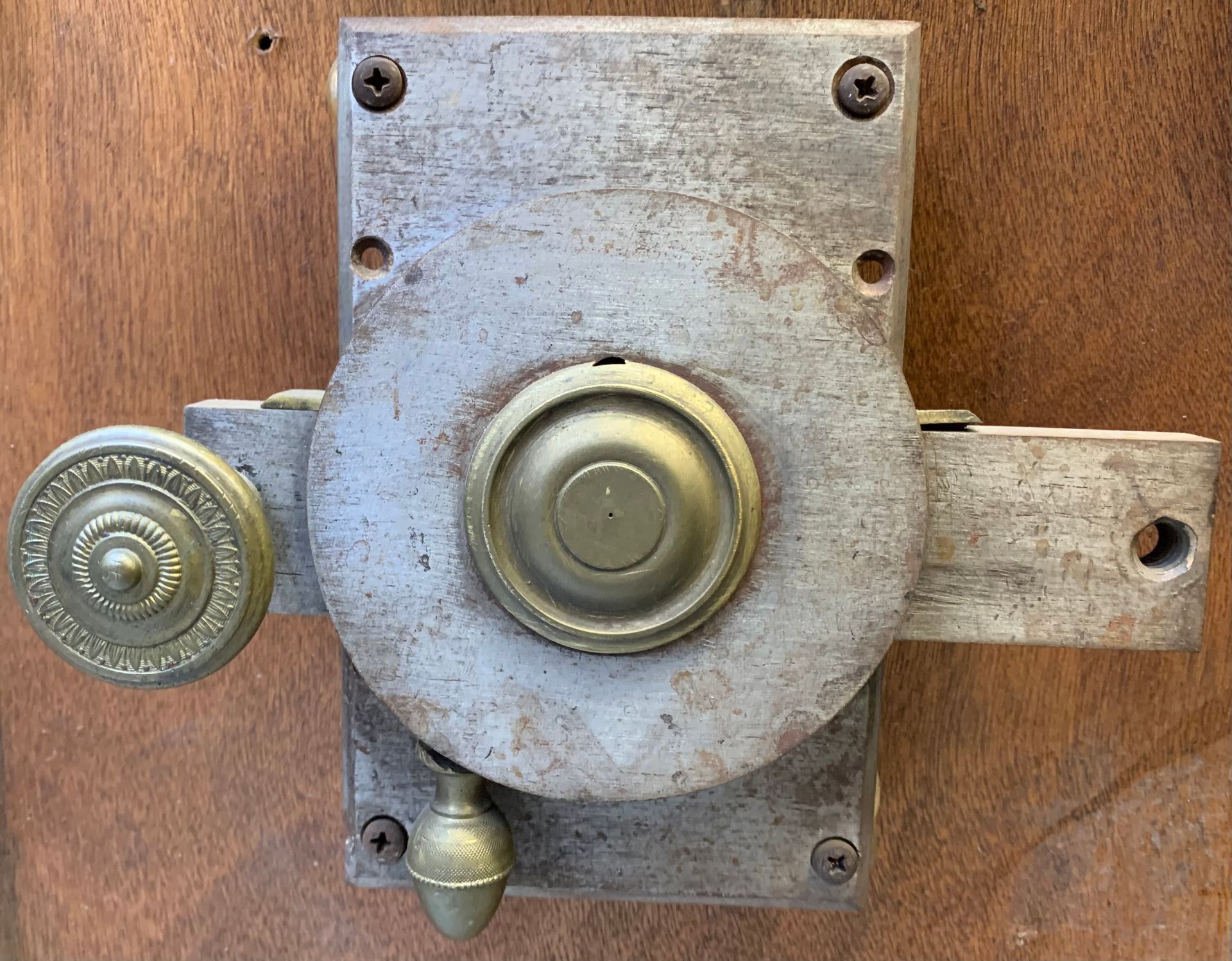 Une serrure à gorge métallique montée sur une porte en bois, comportant un bouton central rond en laiton, une poignée latérale et un petit levier.