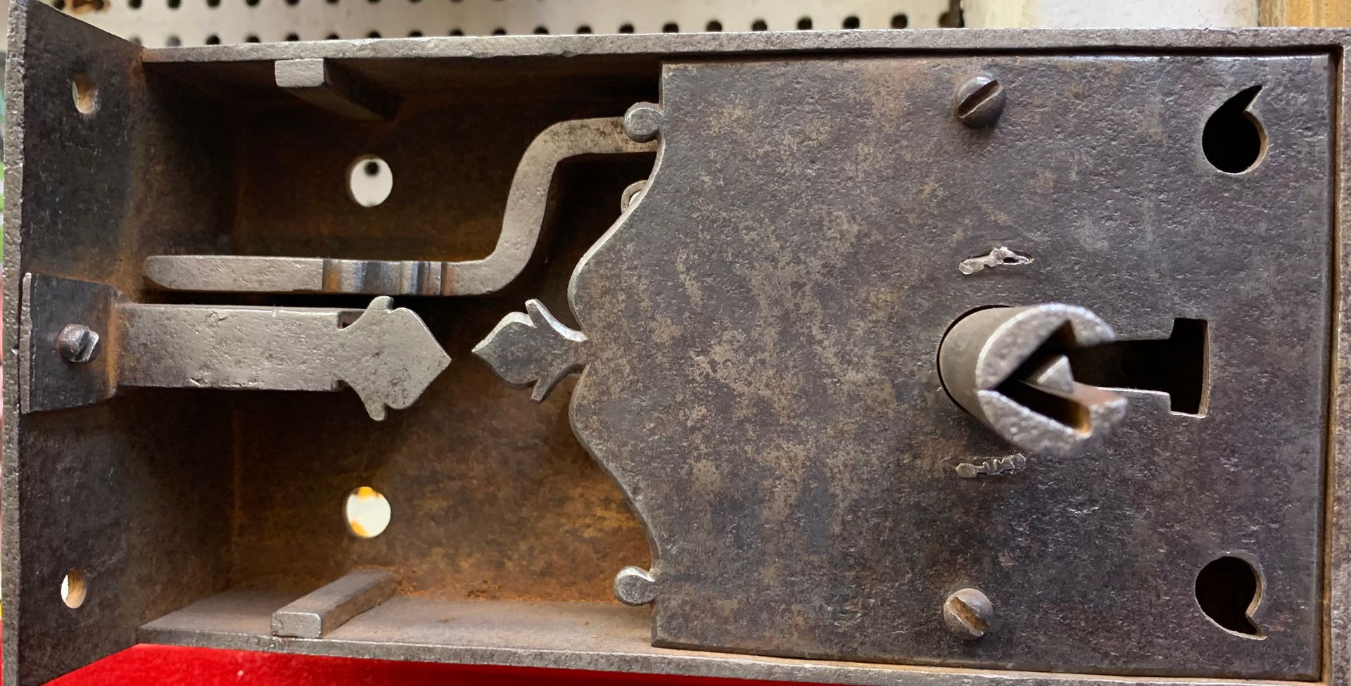 Un mécanisme de serrure à mortaise ancien, ouvert et rouillé, comportant un verrou central et une plaque à trou de serrure aux bords décoratifs.