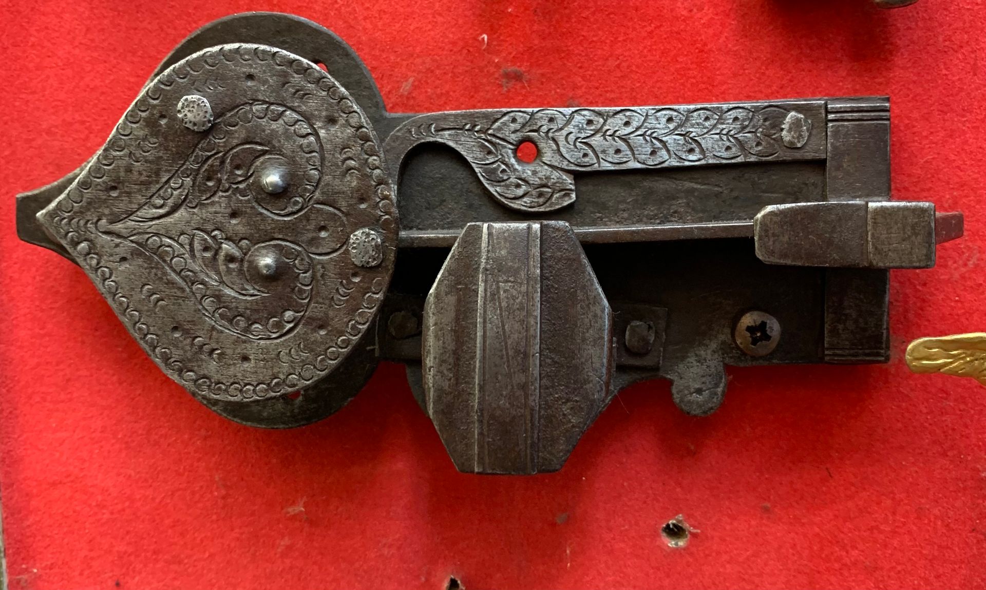 Un loquet ancien en fer forgé, orné d'une plaque en forme de cœur aux gravures florales complexes, sur fond rouge.