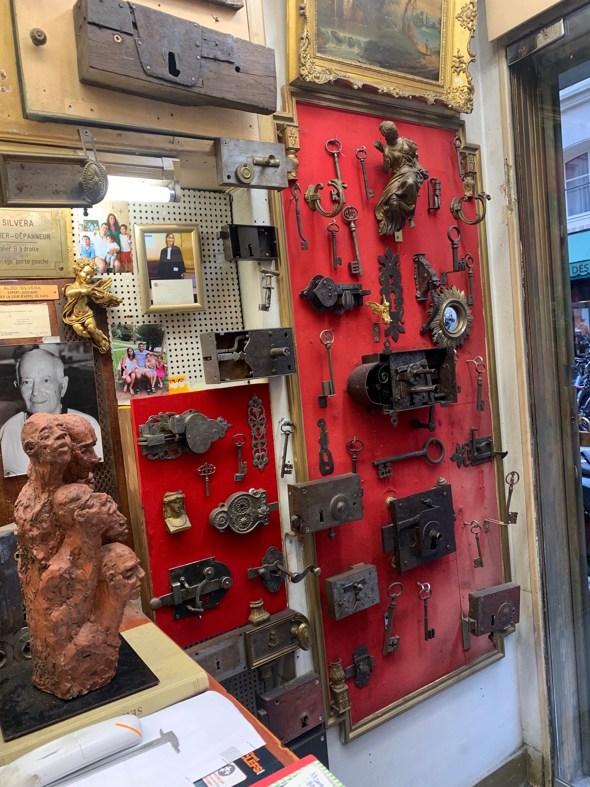 Dans une boutique, une collection de serrures et de clés anciennes est exposée sur des panneaux de velours rouge, avec une figure sculptée au premier plan.