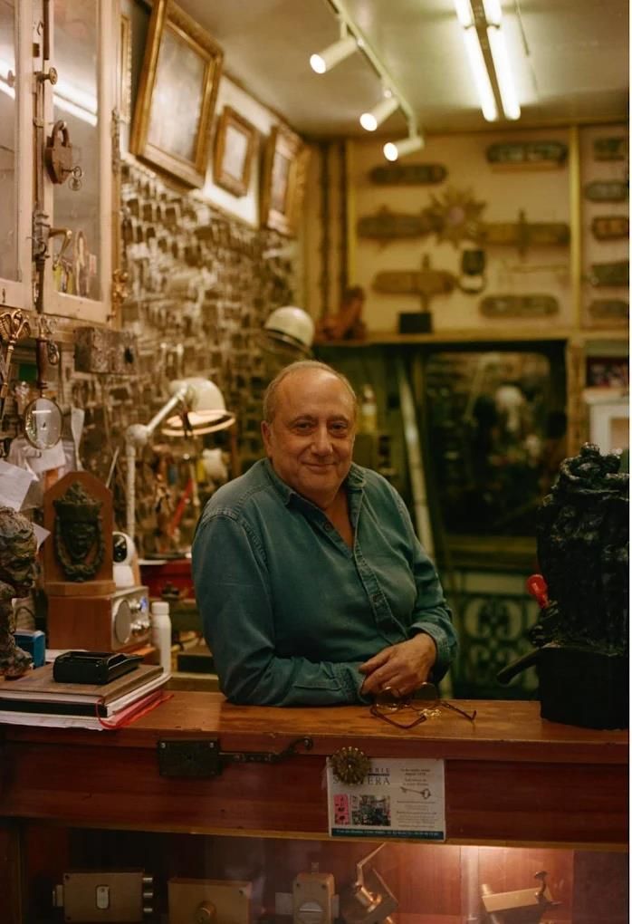 Une personne se tient derrière un comptoir dans une boutique remplie d'objets anciens : quincaillerie, clés et sculptures exposées.