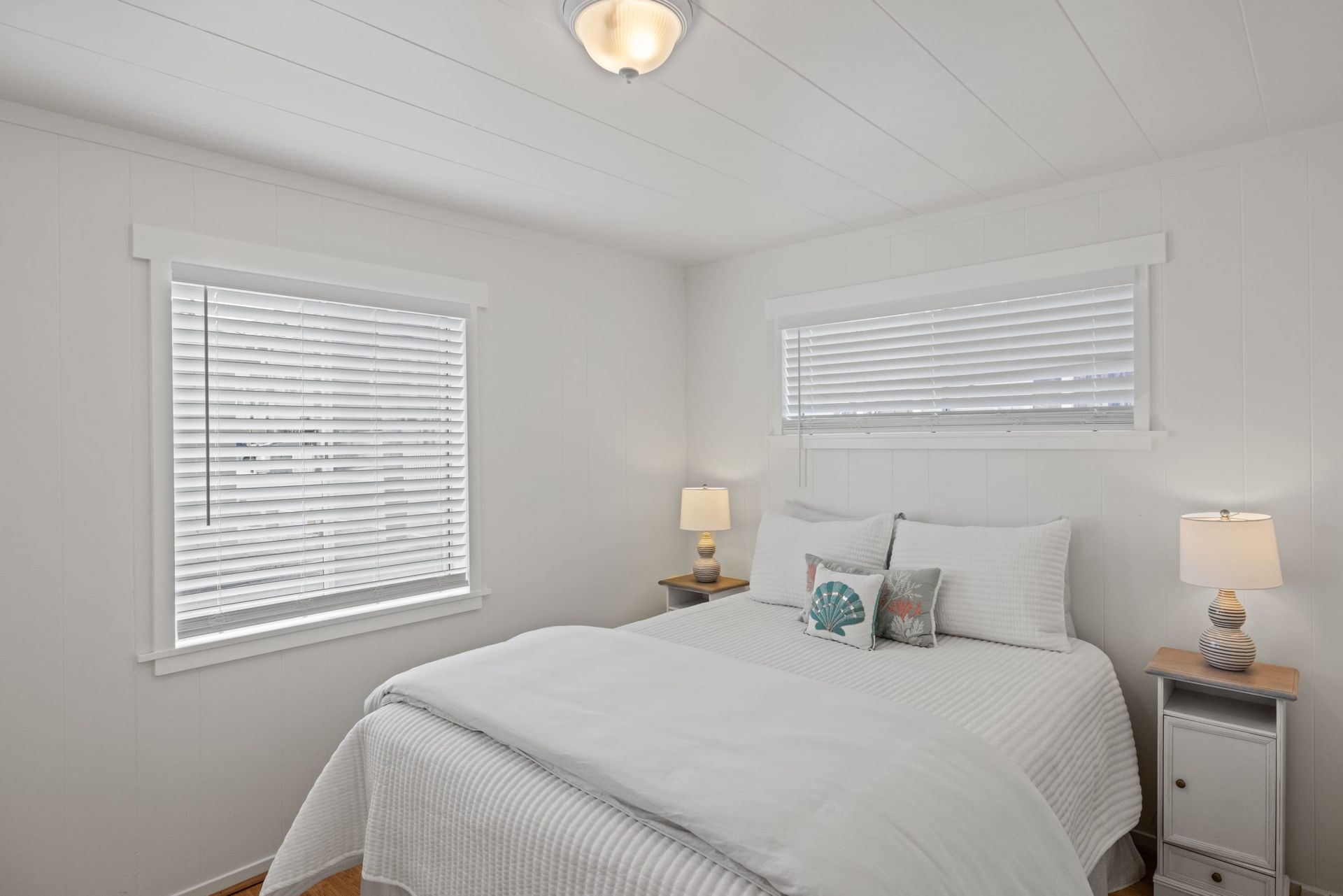A bedroom with a king size bed , two windows , two lamps , and a ceiling fan.